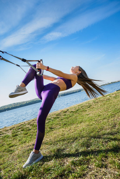 Joven mujer delgada haciendo entrenamiento de suspensión con correas de fitness al aire libre cerca del lago durante el día. Estilo de vida saludable - Foto, imagen