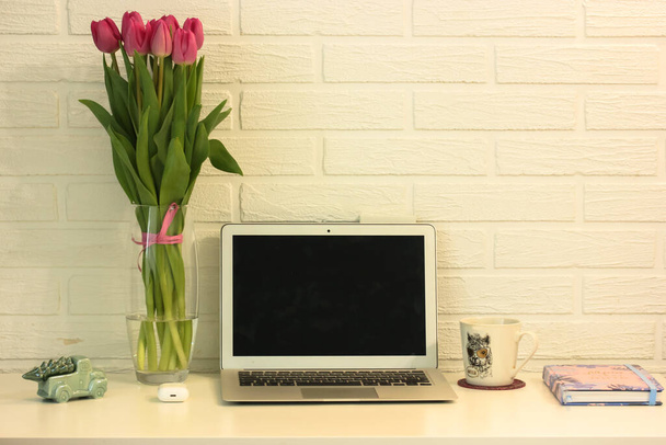 Красиве і затишне місце для дівчини, щоб працювати віддалено. Білий стіл, ноутбук, ваза з рожевими тюльпанами, блокнот і біла чашка порцеляни. Офіс вдома
. - Фото, зображення