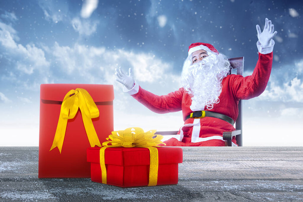 Asian man in Santa costume sitting on a wooden chair with a gift box on the table with snowfall background. Merry Christmas - 写真・画像