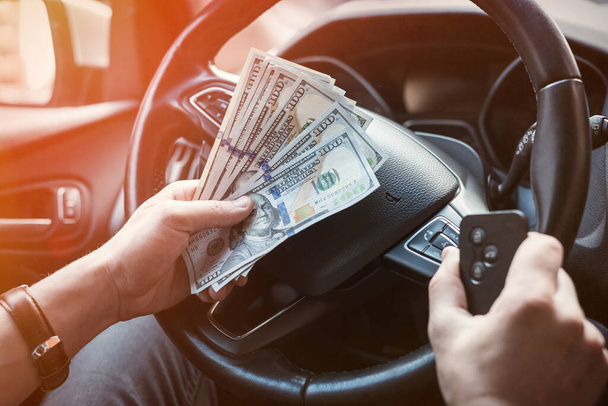 Δολάριο και κλειδί αυτοκινήτου στο αντρικό χέρι μέσα στο αυτοκίνητο. πληρώνουν σε αγαθά ή ενοικιάσεις αυτοκινήτων - Φωτογραφία, εικόνα