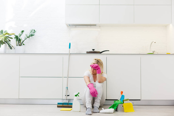 Zmęczona młoda sprzątaczka odpoczywająca w kuchni po prowadzeniu domu.Siedząca na blatach kuchennych i trzymająca mop i spray 'ową butelkę - Zdjęcie, obraz