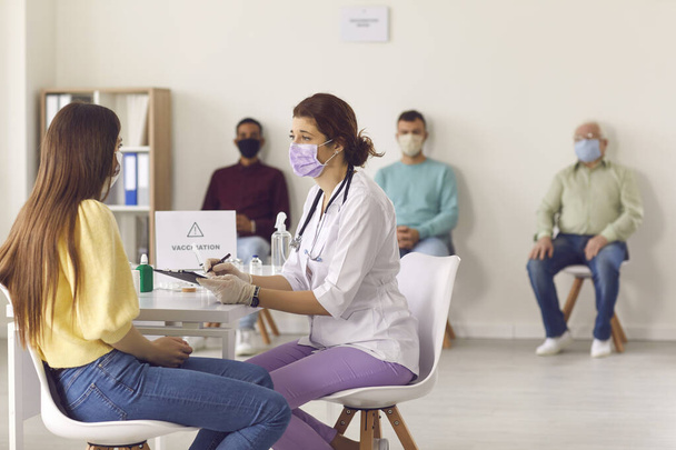 Doctor in face mask interviewing young woman before giving her antiviral vaccine - Photo, image