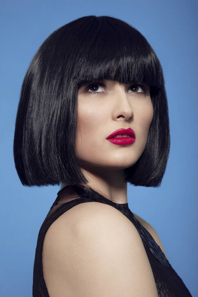 Closeup beauty portrait of a brunette woman in black wig with make up, red lips, over blue background. - Fotoğraf, Görsel