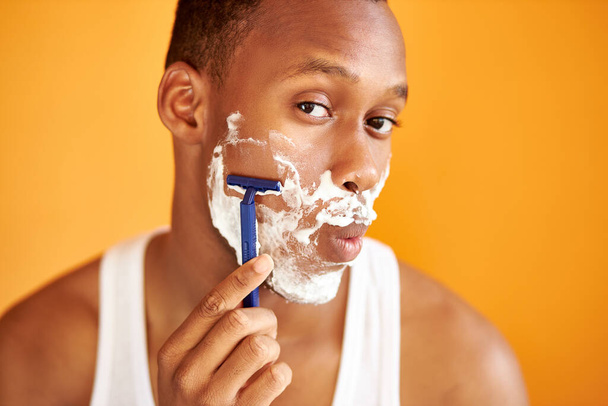 portrait of african male shaving his face with foam and razor isolated on orange background - Fotó, kép