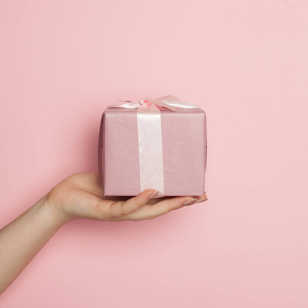 Pink gift box. Minimal background composition with woman hand holding gift on pastel pink color - Fotó, kép