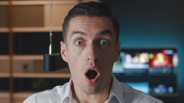 Close-up portret van een bange man in wit shirt uitpuilende ogen en het bedekken van de mond in angst reageren op verschrikkelijke situatie op home office appartement achtergrond in de avond - Video