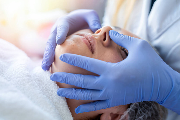 Kosmetologe Hände in blauen Handschuhen berühren Gesicht der jungen Patientin, die Massage macht. Konzept der Hautpflege und Verjüngung. - Foto, Bild