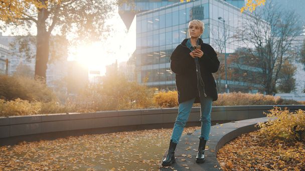 Jovem caucasiana usando telefone no parque da cidade em frente ao prédio de escritórios no outono - Foto, Imagem