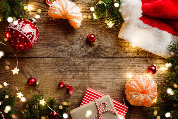 Vrolijke kerstversiering, dennentakken, sinaasappel, geschenkdozen, kerstmuts, lichtslinger, sneeuwvlokken op houten achtergrond. Kerstmis en nieuwjaarsvakantie, bokeh, licht. Vlakke lay, bovenaanzicht - Foto, afbeelding