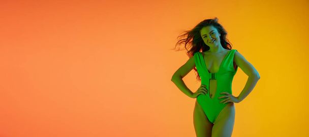 Half-length portrait of beautiful seductive girl in fashionable green swimsuit on bright gradient background in neon light - Photo, Image