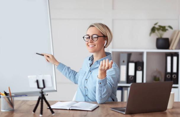 Positive millennial teacher pointing at empty blackboard near laptop at home, giving online lecture, space for design - Photo, Image