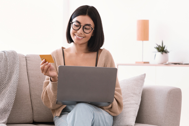 Asian lady in glasses making purchases using laptop holding card - Foto, afbeelding