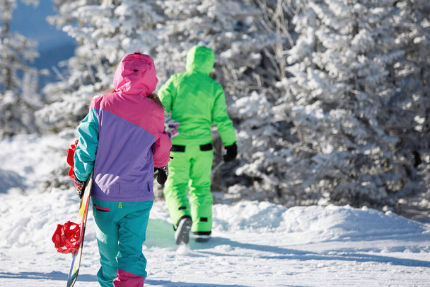 Сноубордисты ходят по пушистому снегу в зимнем хвойном лесу. Они одеты в лыжную одежду: комбинезон с капюшонами. Вид сзади. Здоровый образ жизни. Спортивная концепция. Селективный фокус. - Фото, изображение