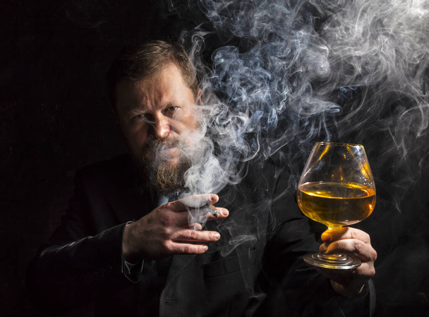 Solide selbstbewusste bärtige Mann im Anzug mit Glas Whisky und Zigarre mit Rauch - Foto, Bild