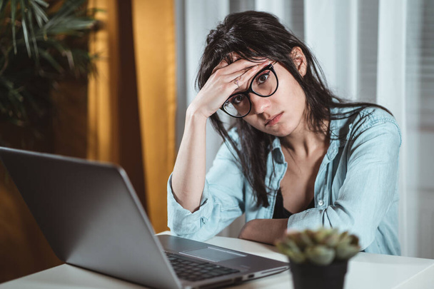 Worried young woman sitting at home desk with laptop, thinking of problems. Pensive unmotivated lady looking away, feeling lack of energy, doing remote freelance tasks at home. Overtime work concept - 写真・画像