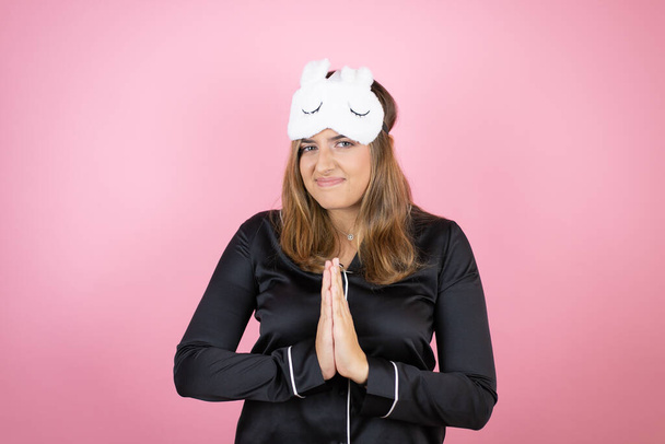 Young caucasian woman wearing sleep mask and pajamas over isolated pink background begging and praying with hands together with hope expression on face very emotional and worried - Photo, Image