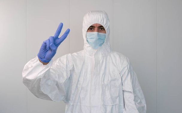 Een mannelijke verpleegster in een beschermend pak en masker die het overwinningsgebaar maakt. Concept van het overwinnen van de Covid-19. Geheide vingers die V-teken vormen, succesvolle overleving van coronavirus patiënten. - Foto, afbeelding