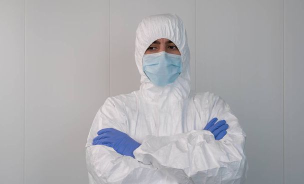 Während der Pandemie, die durch das Covid 19, das Coronavirus, ausgelöst wurde, verschränkt ein Mitarbeiter des Gesundheitswesens in einem Schutzanzug im Krankenhaus die Arme. Der Krankenpfleger trägt eine OP-Maske. - Foto, Bild
