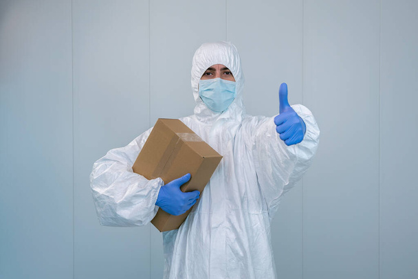 Μια νοσοκόμα με προστατευτική στολή που κούνησε τους αντίχειρες αφού έλαβε ένα κουτί ιατρικών προμηθειών κατά τη διάρκεια της πανδημίας του κορωναϊού, covid 19. Εργάτης υγείας μέσα σε νοσοκομείο με ΜΑΠ και χειρουργική μάσκα. - Φωτογραφία, εικόνα