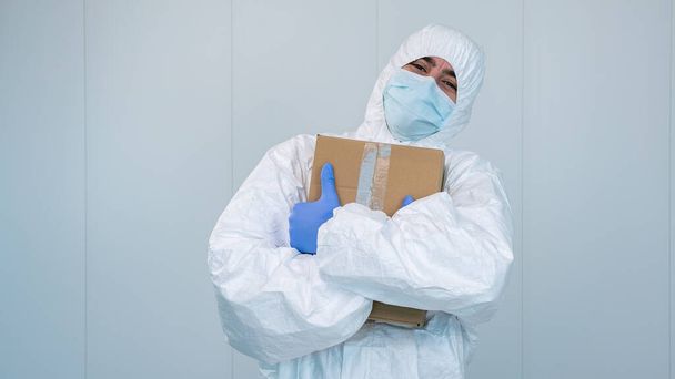 Ένας ευτυχισμένος νοσοκόμος με προστατευτική στολή αγκαλιάζει ένα κουτί με ιατρικές προμήθειες με τα χέρια του στο νοσοκομείο κατά τη διάρκεια πανδημίας που προκλήθηκε από τον ιό Covid 19, τον Coronavirus. Η αρσενική νοσοκόμα φοράει χειρουργική μάσκα.. - Φωτογραφία, εικόνα