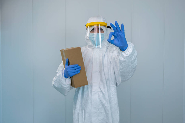 保護服と顔の盾の看護師は、コロナウイルスのパンデミックの間に医療用品箱を受け取った後に親指をジェスチャーで、 coid19 。PPEと外科マスクを持つ病院内の医療従事者 - 写真・画像