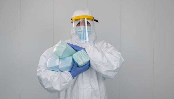 Un operatore sanitario in tuta protettiva PPE porta tra le braccia diverse confezioni di nuove maschere. Il medico indossa una maschera per prevenire il coronavirus. - Foto, immagini