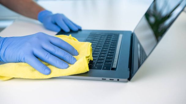 Een gezondheidswerker veegt zijn laptop af met een doek om de verspreiding van het coronavirus te voorkomen. Draagt handschoenen voor het reinigen en desinfecteren van het toetsenbord. - Foto, afbeelding