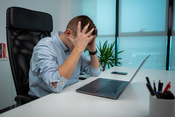 Tired or stressed young businessman sitting in front of computer in office. Unhappy entrepreneur at workplace. Unhealthy exhausted caucasian man having painful feelings overwhelmed with laptop work. - Photo, image