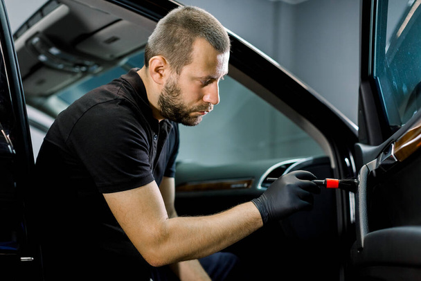 Handsome young Caucasian man, worker of professional car detailing workshop, cleaning car door interior by a soft brush. Car cleaning and detailing concept - 写真・画像