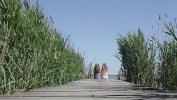 Dolly schot volgen op van twee meisjes zitten met de rug op een houten pier - Video