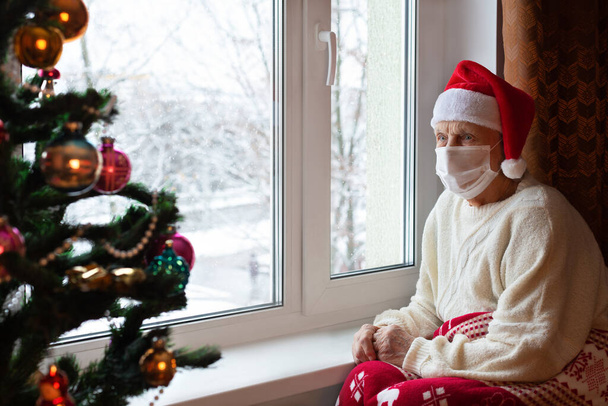 Elderly Caucasian woman wearing Santa hat sits by window wearing mask on her face and sadly looks Christmas tree. Concept: lonely elderly people in quarantine during coronavirus on eve of Christmas holidays. Portrait.Close-up - Foto, Imagem