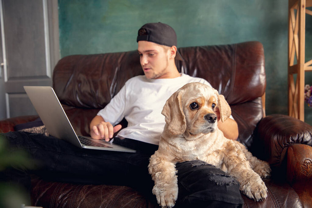 Mladý pohledný muž sedí a pracuje doma se svým roztomilým psem. Útulné kancelářské pracoviště, vzdálená práce, koncepce online učení. - Fotografie, Obrázek