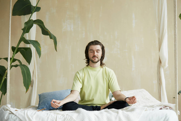 hombre guapo con pelo largo meditando en la cama, hombre sosteniendo brazos en yoga mudra y sentado en pose de loto en casa - Foto, imagen