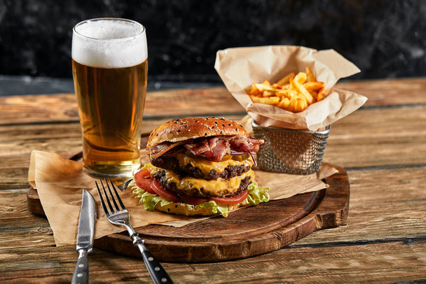 A large mouth-watering burger with grilled beef patty and fresh vegetables. Tasty american cheeseburger on a wooden board. Classic homemade burger in craft paper. - Foto, afbeelding