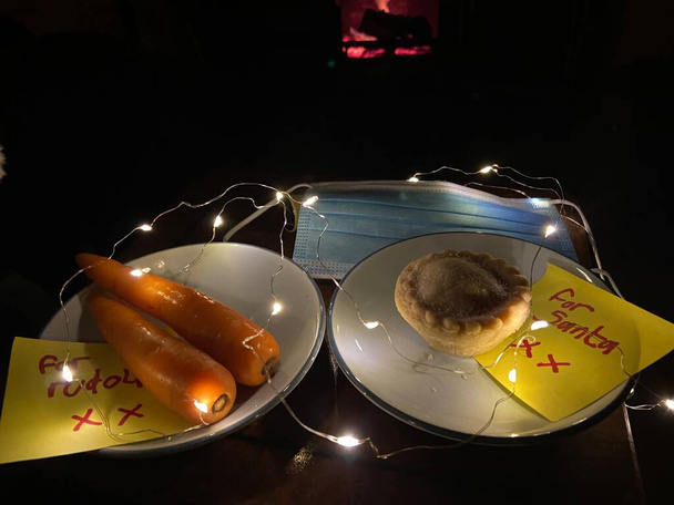 covid-19 Heiligabend, Weihnachtsmütze mit Gesichtsmaske und für Weihnachtsmann Notiz mit Hackfleischkuchen und Karotten für Rudolph in Kinderschrift Notiz   - Foto, Bild