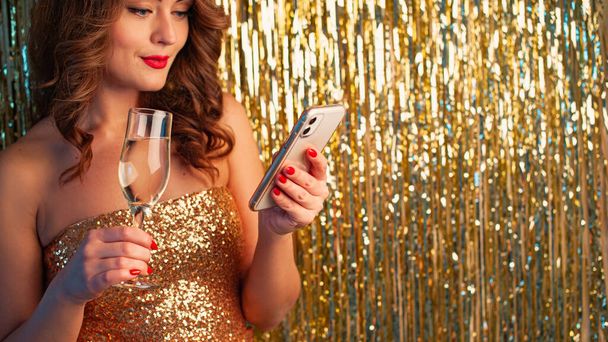 Mujer joven bebe champán, sosteniendo un smartphonee, divirtiéndose sobre un fondo dorado brillante - Foto, imagen