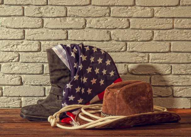 traditional wide-brimmed cowboy hat, rough lasso boots and a striped USA flag on a brick background - Zdjęcie, obraz