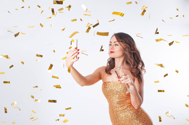 Молодая женщина пьет шампанское, делает селфи на смартфоне, развлекается с золотыми конфетти - Фото, изображение