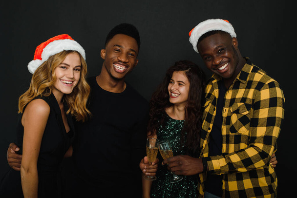 Рождественская вечеринка. Талия до четырех друзей в Санта-Шляпы весело и пить шампанское во время новогодней вечеринки, изолированные на черном фоне - Фото, изображение