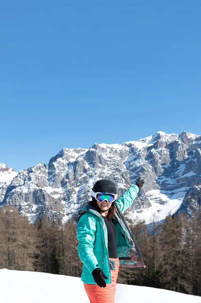Une femme vêtue d'une combinaison de ski colorée, d'un casque et de lunettes de soleil après le ski se dresse sur fond de sommets montagneux. Concept sportif, personnes, voyage - Photo, image