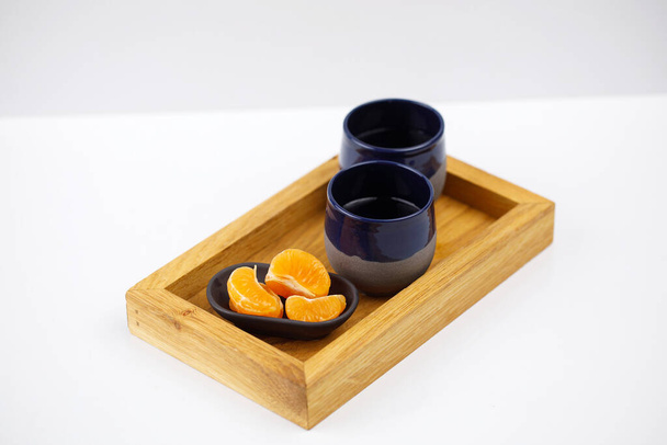 Деревянный поднос с двумя глиняными чашками рядом с подставкой с очищенными апельсиновыми фруктами, все на белом фоне - Фото, изображение