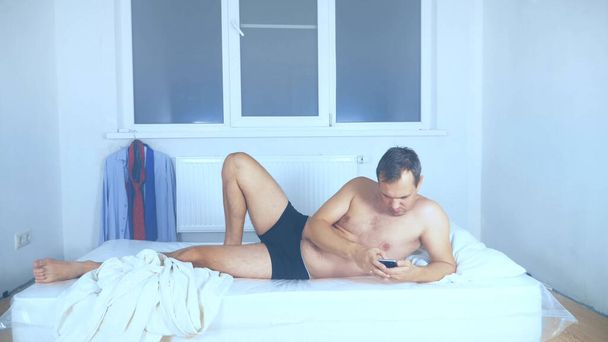 A naked man lies on a mattress in an empty room, using his phone - Foto, afbeelding