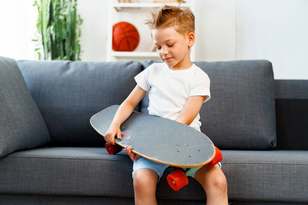 Little boy sitting on sofa at home and holding skateboard - Foto, Imagen