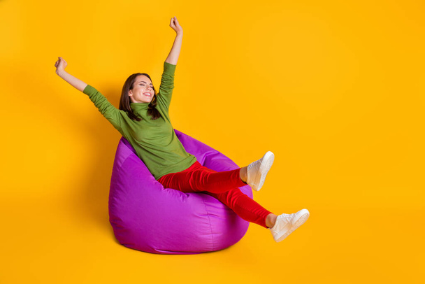 Attractive lady sit comfortable bean bag raise hands legs wear green sweater red pants isolated yellow color background - Foto, imagen