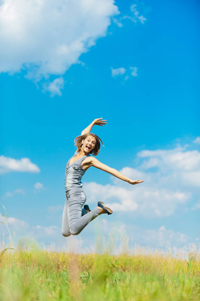 Joven hermosa mujer en un mono de mezclilla azul diversión salta contra el cielo azul con nubes y hierba verde año de altura. - Foto, imagen