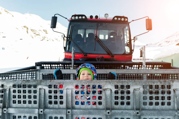 Cute adorable zabawy szczęśliwy maluch chłopiec wewnątrz czerwony nowoczesny snowcat ratrack pług śnieżny box grooming stoi na szczycie alpejski ośrodek narciarski Ischgl Austria. Ciężkie maszyny pojazdy gąsienicowe - Zdjęcie, obraz