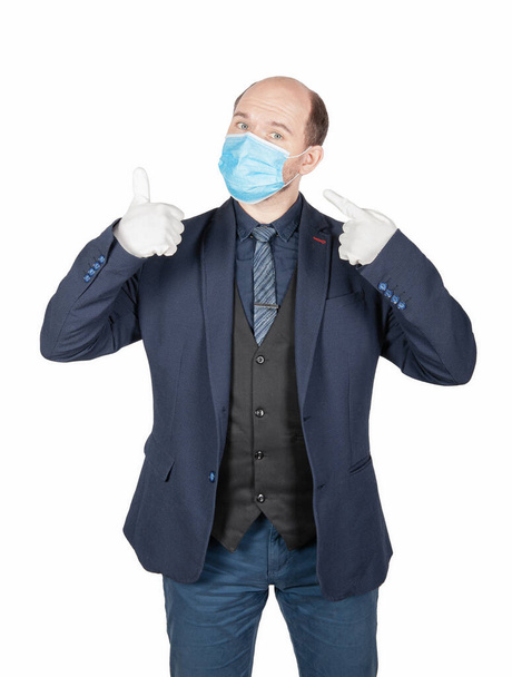 Handsome young business man holding surgical medical virus protection mask with white gloves showing thumbs up isolated on whit - Photo, image