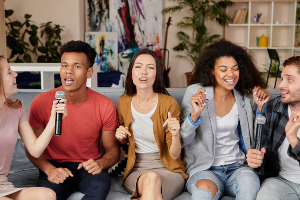 Muzyka przynosi radość. Entuzjastyczne wielokulturowych przyjaciół śpiewających z mikrofonem podczas gry w karaoke w domu, siedzi na kanapie w nowoczesnym mieszkaniu - Zdjęcie, obraz