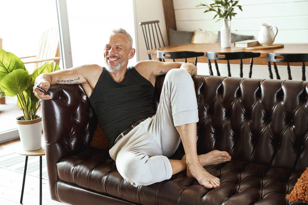 Beau homme d'âge moyen heureux en vêtements décontractés avec smartphone à la main relaxant sur un canapé confortable dans son appartement moderne, regardant de côté et souriant - Photo, image