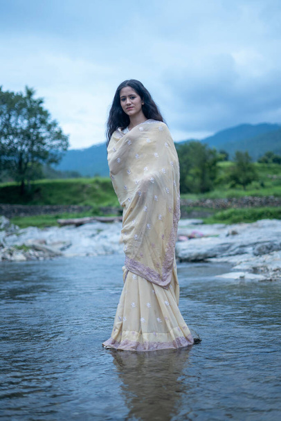 Beautiful Indian young female model wearing traditional saree standing in front of a river landscape with overcast clouds. - Foto, Imagem